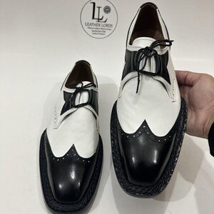 May include: A pair of two-tone wingtip oxford shoes featuring white leather uppers with black leather toe caps and accents. The shoes have black laces and a thick, textured black braided welt. A logo with the text LEATHER LORDS is visible.