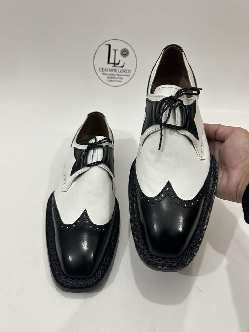 May include: A pair of two-tone wingtip oxford shoes featuring white leather uppers with black leather toe caps and accents. The shoes have black laces and a thick, textured black braided welt. A logo with the text LEATHER LORDS is visible.