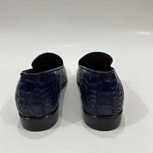 May include: A pair of dark blue loafers featuring a textured crocodile-embossed pattern on the exterior. The shoes have a low-cut silhouette, a rounded heel counter, and a solid black stacked heel, showcasing the back view of the footwear.