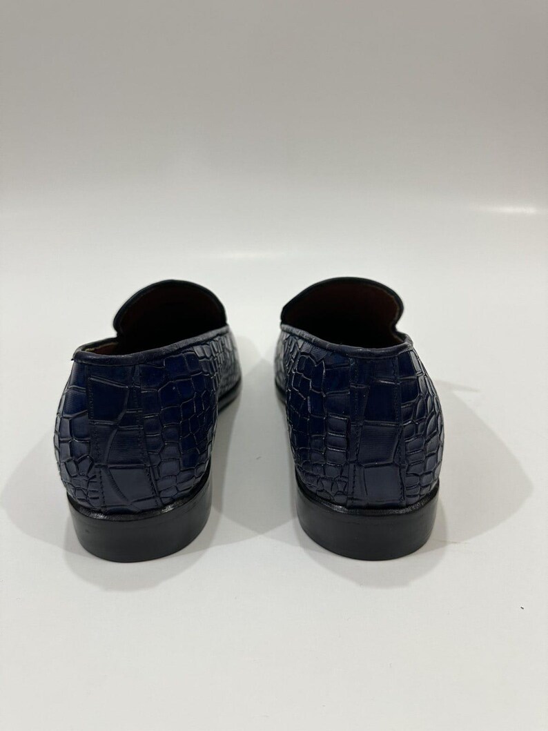 May include: A pair of dark blue loafers featuring a textured crocodile-embossed pattern on the exterior. The shoes have a low-cut silhouette, a rounded heel counter, and a solid black stacked heel, showcasing the back view of the footwear.