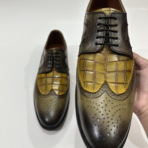 Puede incluir: Un par de zapatos oxford de cuero con dise&ntilde;o bicolor, con puntera y tal&oacute;n en verde oliva oscuro y empeine de cuero con relieve de cocodrilo en color amarillo mostaza. Incluyen cordones negros y detalles de estilo brogue.