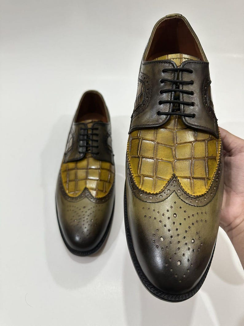 Puede incluir: Un par de zapatos oxford de cuero con dise&ntilde;o bicolor, con puntera y tal&oacute;n en verde oliva oscuro y empeine de cuero con relieve de cocodrilo en color amarillo mostaza. Incluyen cordones negros y detalles de estilo brogue.