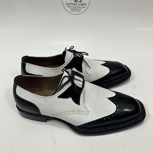 May include: A pair of black and white leather wingtip oxford shoes featuring a two-tone design, black laces, and decorative brogue detailing along the toe cap and sides. The shoes are displayed below a circular logo reading LEATHER LORDS.