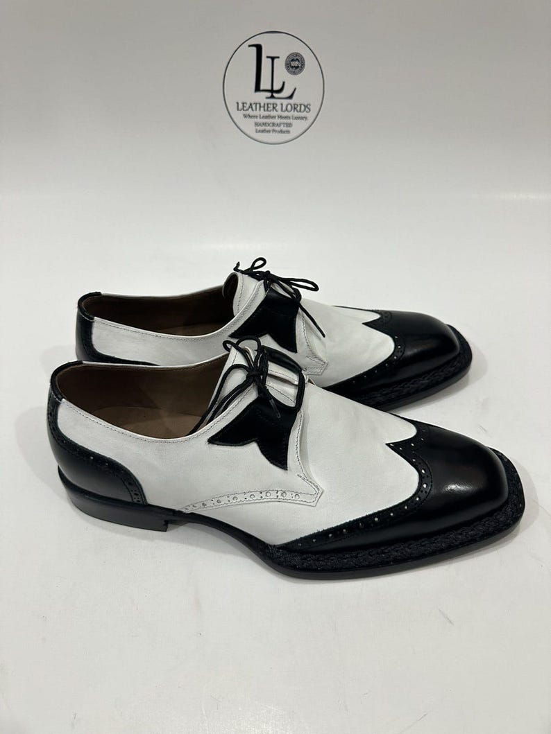 May include: A pair of black and white leather wingtip oxford shoes featuring a two-tone design, black laces, and decorative brogue detailing along the toe cap and sides. The shoes are displayed below a circular logo reading LEATHER LORDS.