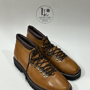 May include: A pair of tan leather hiking boots with dark brown padded collars, dark brown laces, and metal D-ring eyelets. The boots feature a thick black lugged rubber sole with white stitching along the welt. A logo reads LEATHER LORDS.
