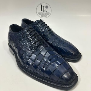 May include: A pair of dark navy blue lace-up dress shoes featuring a textured crocodile skin pattern, black laces, and a braided trim along the sole. The logo for Leather Lords is visible in the background above the shoes.