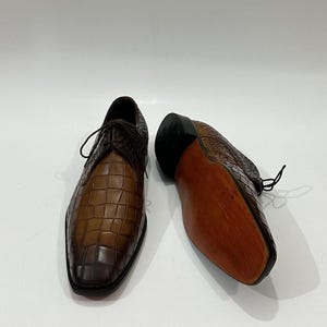 May include: A pair of brown leather dress shoes featuring a textured crocodile-embossed pattern. One shoe is positioned upright showing the lace-up front, while the other is placed on its side to reveal a smooth, light brown wooden sole and heel.