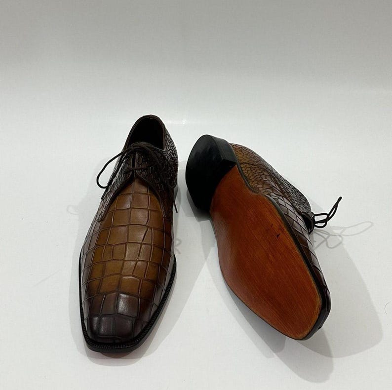 May include: A pair of brown leather dress shoes featuring a textured crocodile-embossed pattern. One shoe is positioned upright showing the lace-up front, while the other is placed on its side to reveal a smooth, light brown wooden sole and heel.