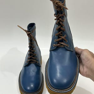 以下が含まれることがあります： 茶色のレザーシューレース、銀色の金属製アイレット、厚みのある質感のタンカラーのラバーラグソールを備えた青いレザーのレースアップブーツ。ブーツは滑らかな仕上げで、ソールのウェルトに沿って白いステッチが見えます。
