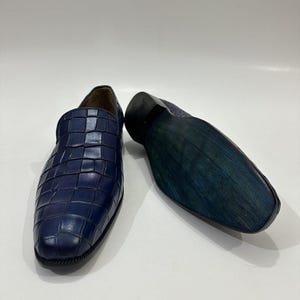 May include: A pair of navy blue loafers featuring a textured crocodile-embossed pattern on the upper. One shoe is positioned upright to show the front, while the other is placed on its side to reveal the dark blue, smooth, flat sole.