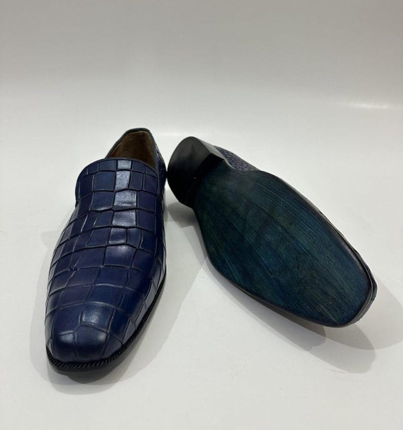 May include: A pair of navy blue loafers featuring a textured crocodile-embossed pattern on the upper. One shoe is positioned upright to show the front, while the other is placed on its side to reveal the dark blue, smooth, flat sole.
