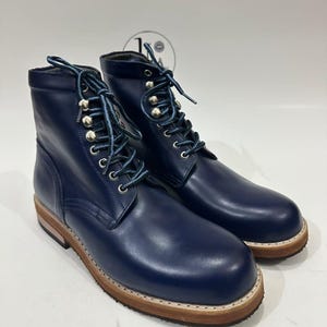 Puede incluir: Un par de botas de cuero azul marino con cordones frontales, ojales y ganchos de metal plateado. Las botas tienen cordones con estampado azul y blanco, una entresuela de madera clara apilada y una suela exterior de color marrón oscuro.
