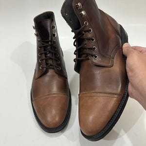 May include: A pair of brown leather lace-up boots featuring a cap toe design, dark brown laces, and silver-toned metal eyelets. The boots have a smooth leather finish, a structured ankle-high silhouette, and a thick black rubber sole.