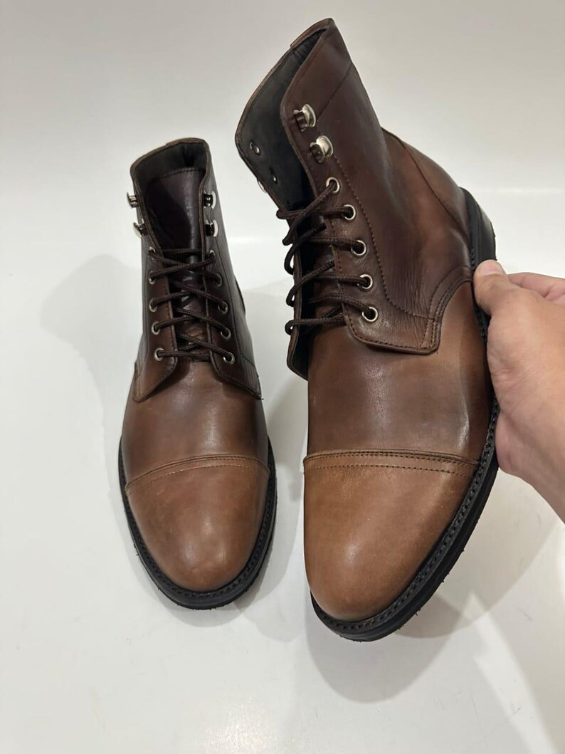 May include: A pair of brown leather lace-up boots featuring a cap toe design, dark brown laces, and silver-toned metal eyelets. The boots have a smooth leather finish, a structured ankle-high silhouette, and a thick black rubber sole.