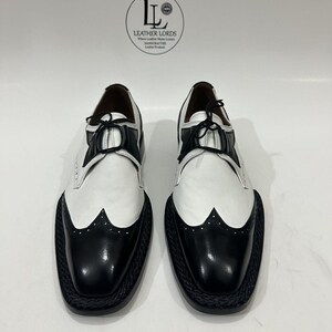 May include: A pair of black and white leather wingtip oxford shoes with black laces and a braided black sole edge. The shoes feature a logo with the text LEATHER LORDS Where Leather Meets Luxury HANDCRAFTED Leather Products.