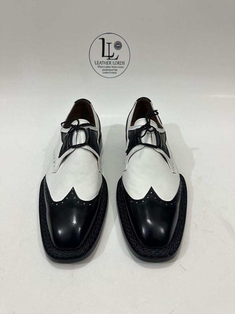 May include: A pair of black and white leather wingtip oxford shoes with black laces and a braided black sole edge. The shoes feature a logo with the text LEATHER LORDS Where Leather Meets Luxury HANDCRAFTED Leather Products.
