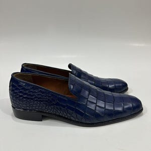 May include: A pair of dark blue slip-on loafers featuring a textured crocodile-embossed pattern across the upper surface. The shoes have a low heel, a rounded toe, and a smooth brown interior lining, presented against a plain white background.