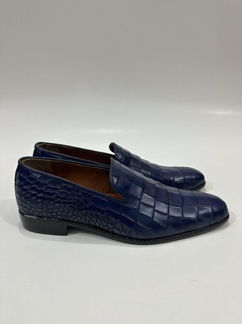 May include: A pair of dark blue slip-on loafers featuring a textured crocodile-embossed pattern across the upper surface. The shoes have a low heel, a rounded toe, and a smooth brown interior lining, presented against a plain white background.