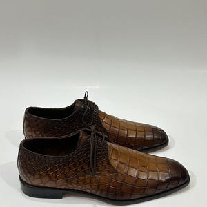 May include: A pair of brown leather lace-up shoes featuring a textured crocodile-embossed pattern. The shoes have a gradient finish, transitioning from a lighter tan in the center to a darker brown at the toe and heel, with thin brown laces.