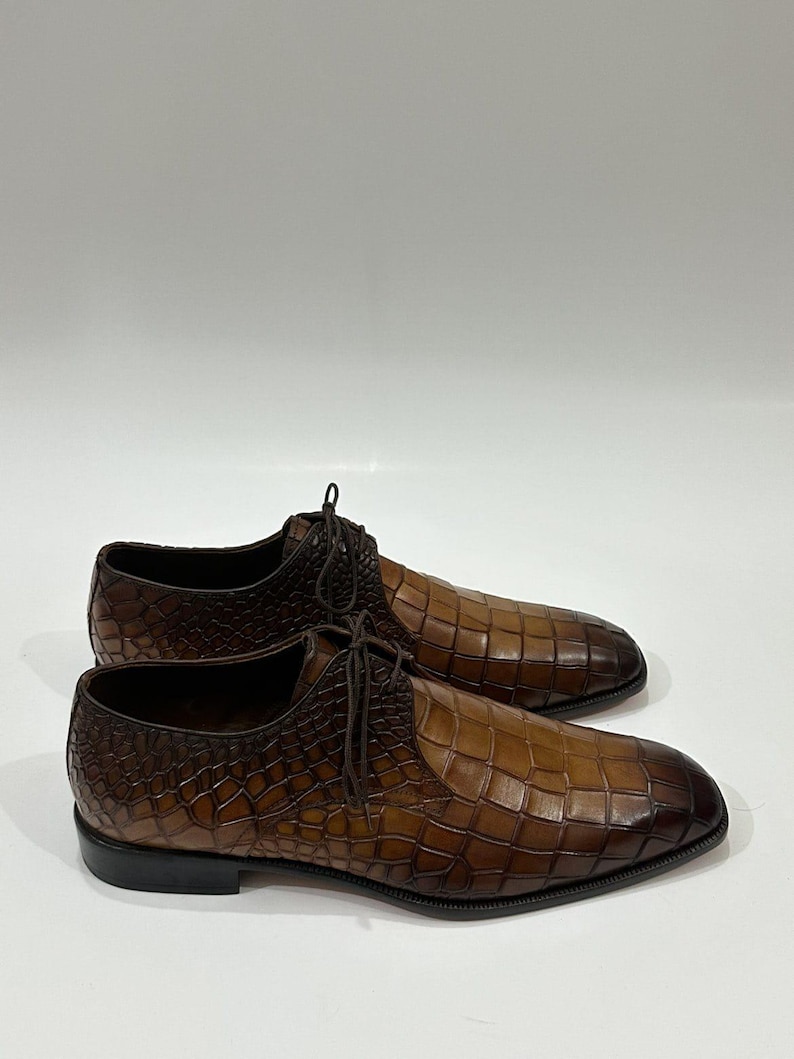 May include: A pair of brown leather lace-up shoes featuring a textured crocodile-embossed pattern. The shoes have a gradient finish, transitioning from a lighter tan in the center to a darker brown at the toe and heel, with thin brown laces.