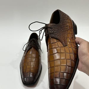 May include: A pair of brown leather dress shoes featuring a prominent crocodile skin texture. The shoes have a gradient color finish, transitioning from a lighter tan in the center to a deep, dark brown at the toe and heel, with dark brown laces.