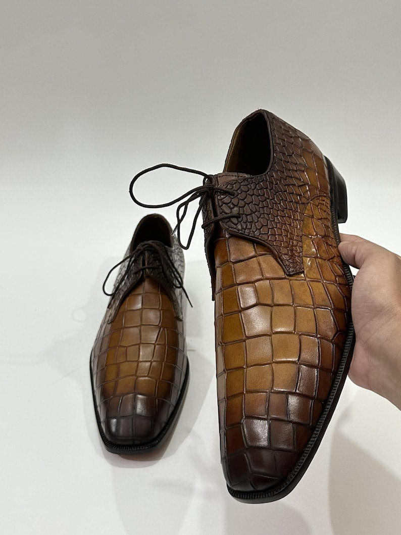 May include: A pair of brown leather dress shoes featuring a prominent crocodile skin texture. The shoes have a gradient color finish, transitioning from a lighter tan in the center to a deep, dark brown at the toe and heel, with dark brown laces.