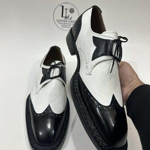 May include: A pair of black and white wingtip shoes featuring a two-tone leather design, black lace-up closures, and a thick black braided trim along the sole edge. The shoes display a classic brogue pattern on the toe cap and heel.