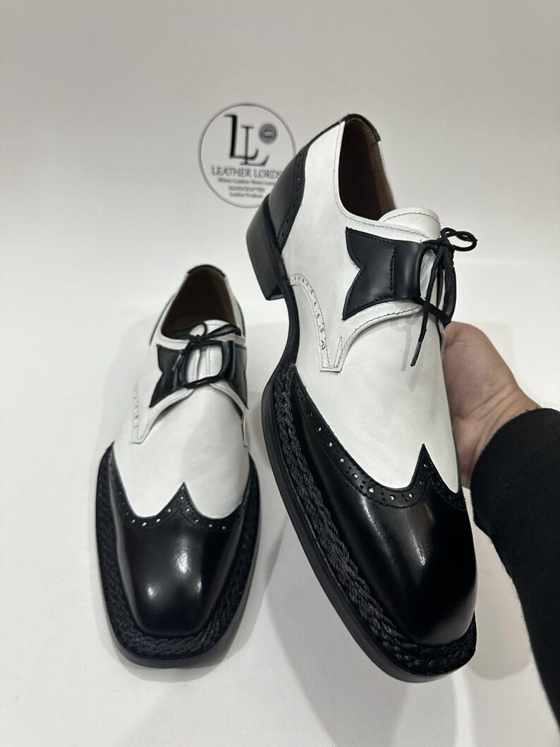 May include: A pair of black and white wingtip shoes featuring a two-tone leather design, black lace-up closures, and a thick black braided trim along the sole edge. The shoes display a classic brogue pattern on the toe cap and heel.