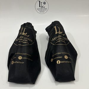 Puede incluir: Dos bolsas de tela negra con cord&oacute;n que presentan el logotipo y el texto dorado Leather Lords Handcrafted Leather Products, junto con iconos de redes sociales e informaci&oacute;n del sitio web. Un logotipo circular se sit&uacute;a sobre las bolsas.