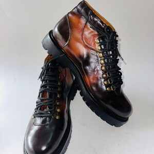 May include: A pair of ankle boots crafted from polished brown leather with a gradient finish. The boots feature black laces, brass-toned metal eyelets, and a thick, black lugged rubber sole. The design includes a rounded toe and a high-top silhouette.