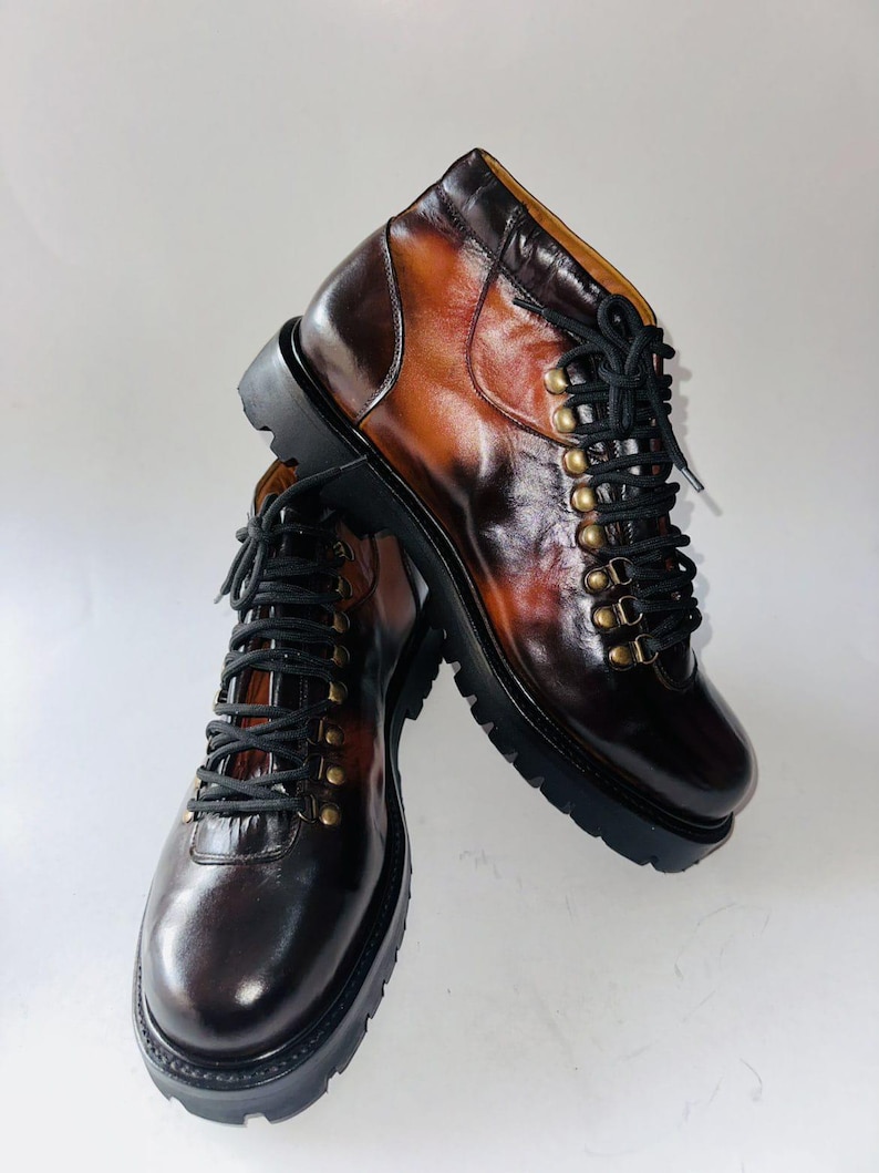 May include: A pair of ankle boots crafted from polished brown leather with a gradient finish. The boots feature black laces, brass-toned metal eyelets, and a thick, black lugged rubber sole. The design includes a rounded toe and a high-top silhouette.