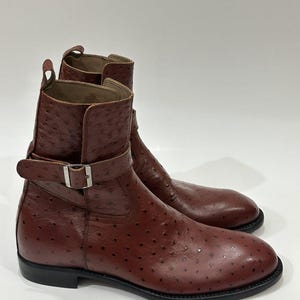 May include: A pair of brown ankle boots featuring a distinct textured quill pattern across the leather surface. Each boot includes a side strap with a silver-toned metal buckle, a pull tab at the top, and a solid black sole with a low heel.