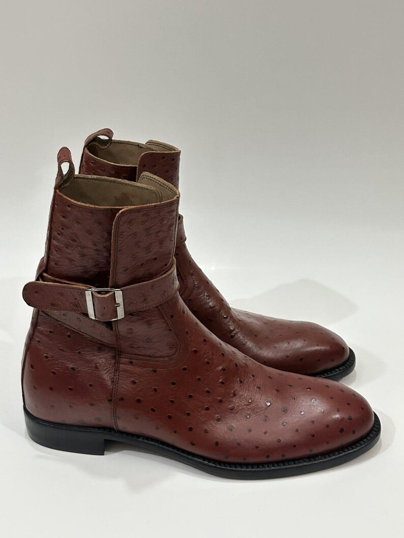 May include: A pair of brown ankle boots featuring a distinct textured quill pattern across the leather surface. Each boot includes a side strap with a silver-toned metal buckle, a pull tab at the top, and a solid black sole with a low heel.