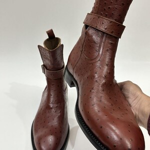 May include: A pair of brown ankle boots featuring a textured ostrich-style leather finish, a rounded toe, a black sole, and a decorative strap with a metal buckle around the ankle. The boots have a pull tab at the top of the shaft.