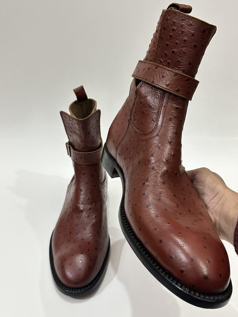 May include: A pair of brown ankle boots featuring a textured ostrich-style leather finish, a rounded toe, a black sole, and a decorative strap with a metal buckle around the ankle. The boots have a pull tab at the top of the shaft.