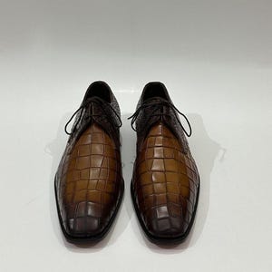 May include: A pair of lace-up shoes featuring a two-tone brown crocodile-embossed leather finish. The shoes have a gradient color effect, transitioning from a lighter tan at the center to a darker brown at the toe and heel areas.