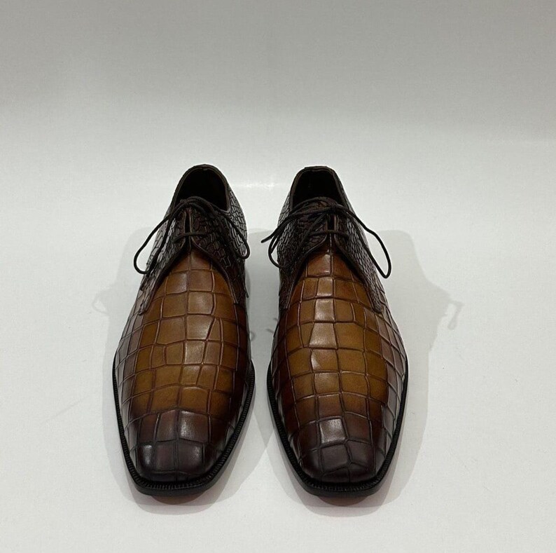 May include: A pair of lace-up shoes featuring a two-tone brown crocodile-embossed leather finish. The shoes have a gradient color effect, transitioning from a lighter tan at the center to a darker brown at the toe and heel areas.