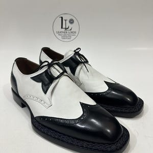 May include: A pair of black and white leather oxford shoes featuring a wingtip design with brogue detailing, black laces, and a braided trim along the sole edge. The shoes have a glossy black toe cap and heel counter with a matte white leather body.
