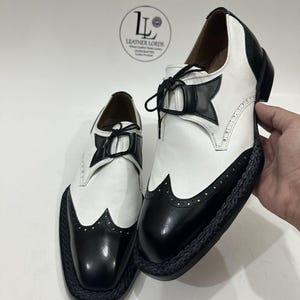 May include: A pair of black and white wingtip oxford shoes featuring a smooth leather upper, black laces, and a braided black trim along the sole edge. The shoes display a classic brogue pattern with decorative perforations on the toe cap.