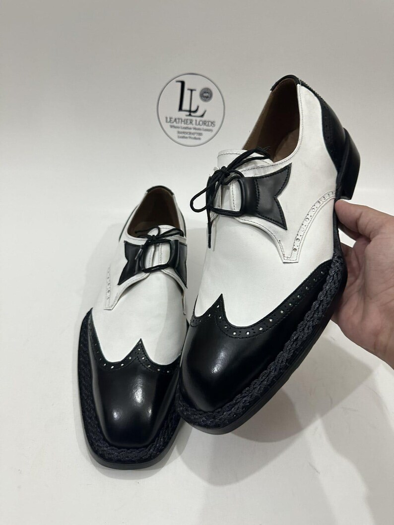 May include: A pair of black and white wingtip oxford shoes featuring a smooth leather upper, black laces, and a braided black trim along the sole edge. The shoes display a classic brogue pattern with decorative perforations on the toe cap.