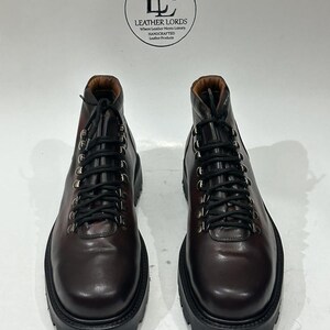 May include: A pair of dark brown leather boots with black laces, silver-toned metal eyelets, and thick black lug soles. A circular logo above the boots reads LEATHER LORDS Where Leather Meets Luxury HANDCRAFTED Leather Products.
