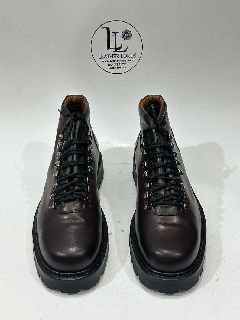 May include: A pair of dark brown leather boots with black laces, silver-toned metal eyelets, and thick black lug soles. A circular logo above the boots reads LEATHER LORDS Where Leather Meets Luxury HANDCRAFTED Leather Products.