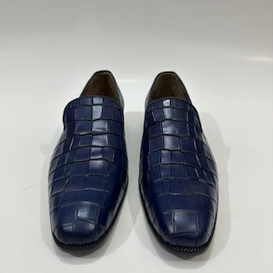 May include: A pair of navy blue slip-on loafers featuring a textured crocodile-embossed pattern across the upper surface. The shoes have a rounded toe, a low profile, and a dark sole with a visible stitched edge detail around the base.