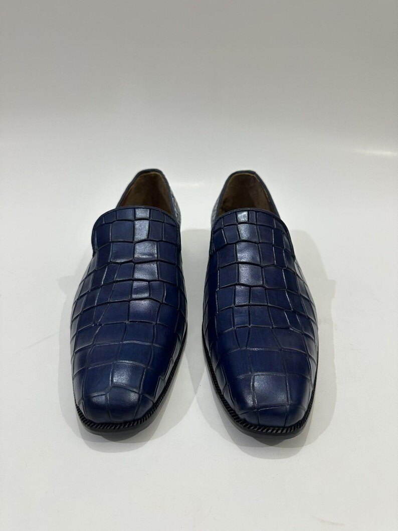 May include: A pair of navy blue slip-on loafers featuring a textured crocodile-embossed pattern across the upper surface. The shoes have a rounded toe, a low profile, and a dark sole with a visible stitched edge detail around the base.