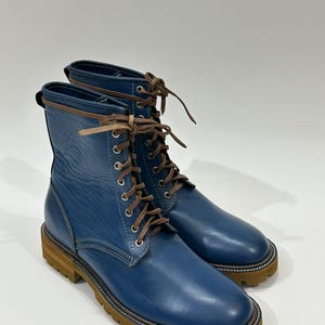 May include: A pair of blue leather lace-up boots with brown laces and a brown sole. The boots have a round toe and a mid-calf height. The boots are on a white background.