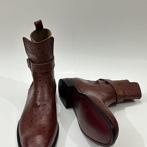 May include: A pair of brown ankle boots featuring a textured ostrich-skin pattern. Each boot has a buckled strap around the ankle, a pull tab at the top, a rounded toe, and a dark sole. The leather has a distinct spotted finish throughout.