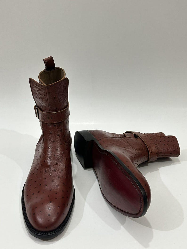 May include: A pair of brown ankle boots featuring a textured ostrich-skin pattern. Each boot has a buckled strap around the ankle, a pull tab at the top, a rounded toe, and a dark sole. The leather has a distinct spotted finish throughout.