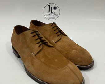 May include: A pair of tan suede leather shoes with dark brown laces and a dark brown sole. The shoes feature a stitched apron toe design and a logo on the background that reads LEATHER LORDS Where Leather Meets Luxury HANDCRAFTED Leather Products.