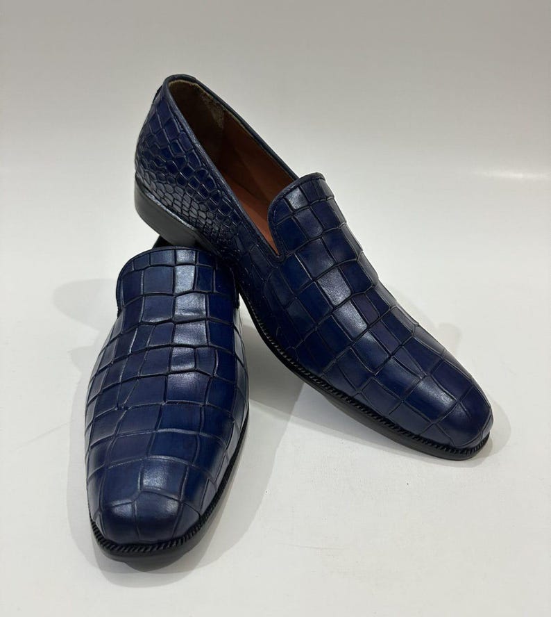 May include: A pair of navy blue slip-on loafers featuring a textured, embossed grid pattern across the upper surface. The shoes have a rounded toe, a low heel, and a smooth interior lining, showcasing a consistent deep blue color throughout.
