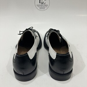 May include: A pair of black and white wingtip oxford shoes featuring a two-tone leather design, black laces, and decorative brogue detailing on the heel counters. The shoes are positioned with the heels facing forward, displaying a stacked heel.